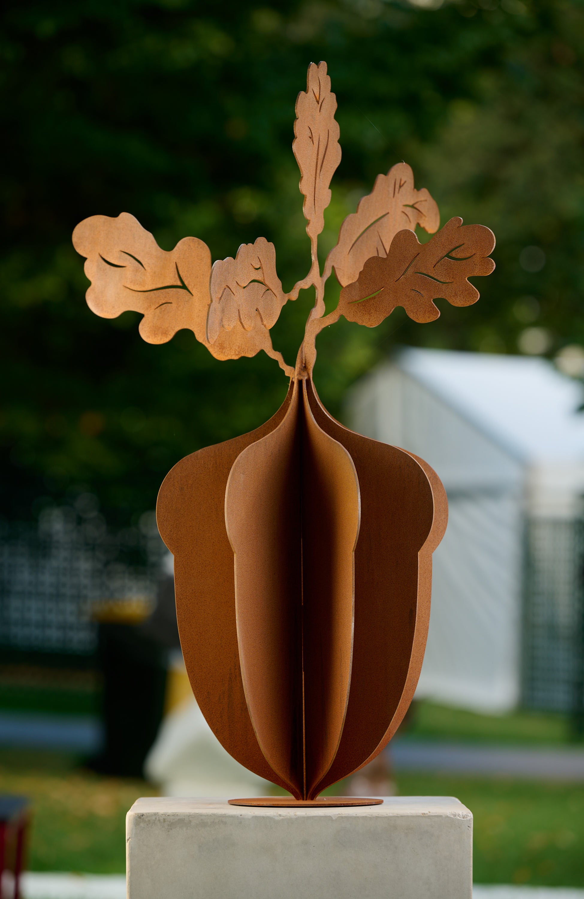 : A Corten rust 3-layer metal sculpture sitting on a concrete display stand under some tall oak trees, featuring an artistic design. Brief Description: This image showcases a Corten rust 3-layer metal sculpture positioned on a concrete display stand. The sculpture is situated under tall oak trees, highlighting its artistic and natural design. The layers and rust finish give it a rustic and elegant appearance, blending harmoniously with the surrounding nature.