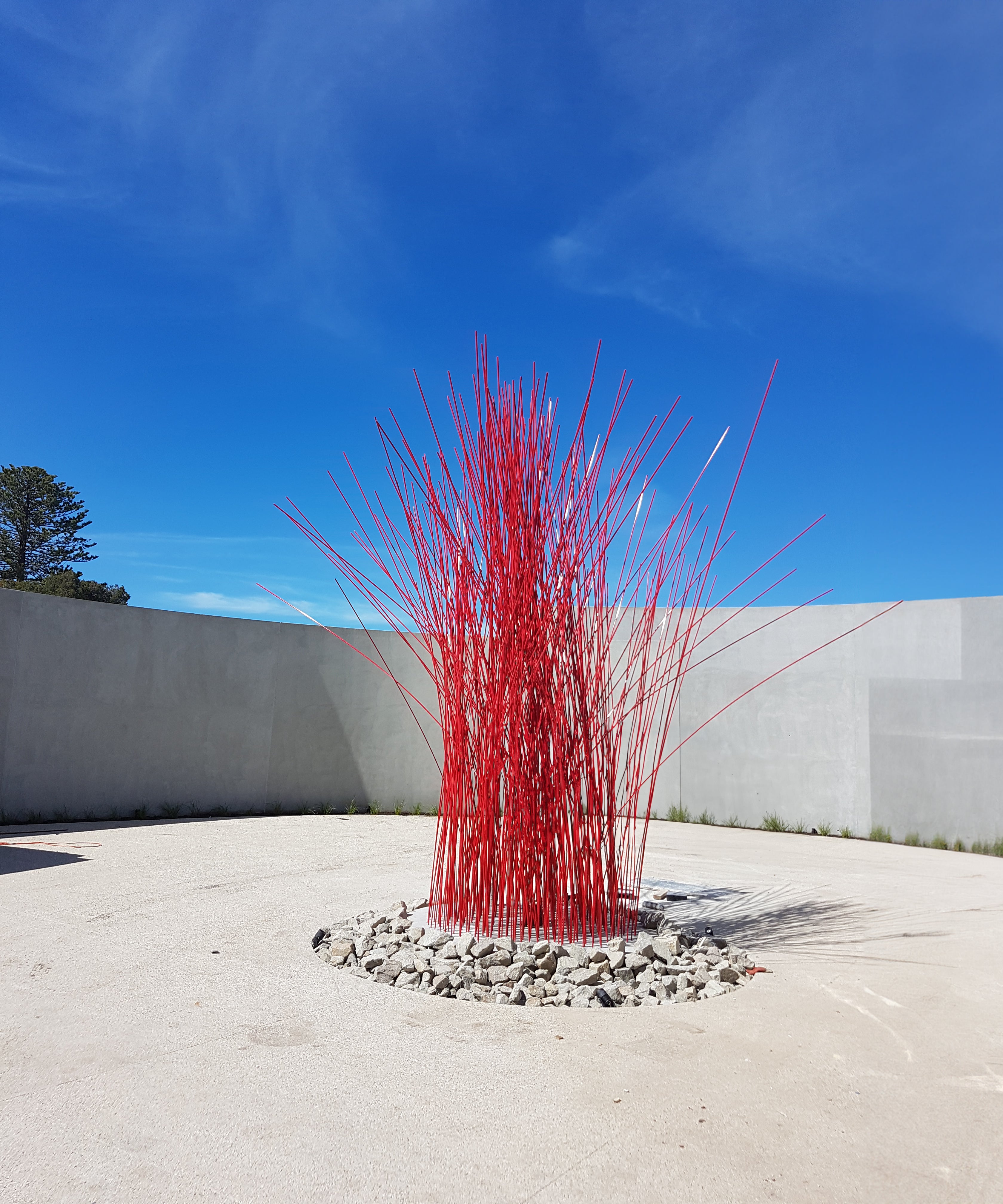 "Large stick sculpture - Entanglements Metal Art Melbourne. A striking red steel sculpture prominently displayed in a concrete wall, creating a captivating focal point in a circular driveway in Mornington."